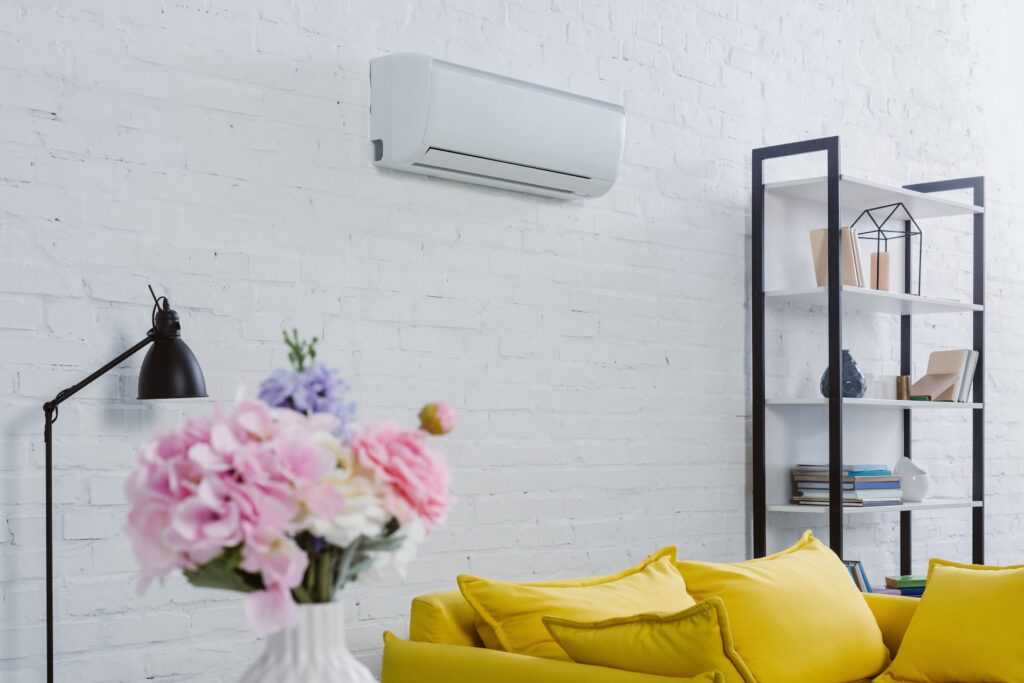 Air conditioning unit mounted on a white wall above a cozy yellow sofa, with a black lamp and decorative bookshelf, illustrating indoor cooling solutions for home comfort.
